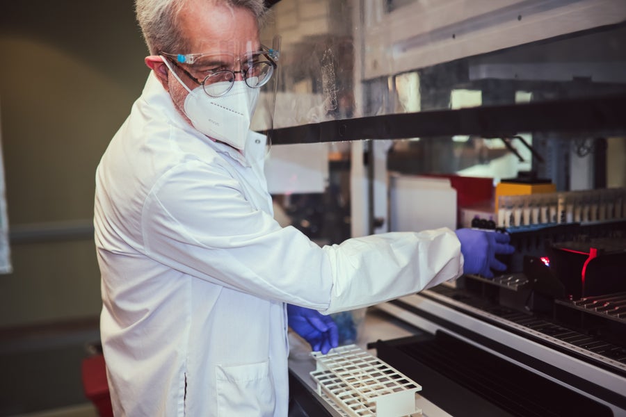 A man in a lab coat and mask stands at a fume hood surrounded by lab equipment
