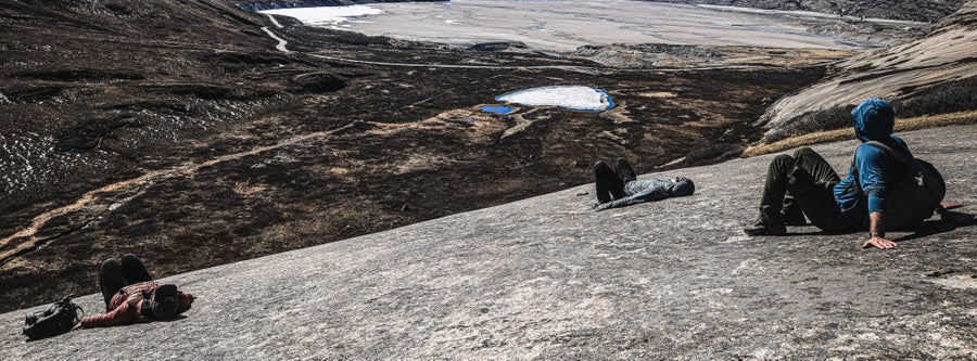 Three people lay or sit on a broad, smooth rock surface that looks out over a glaciated valley and river delta