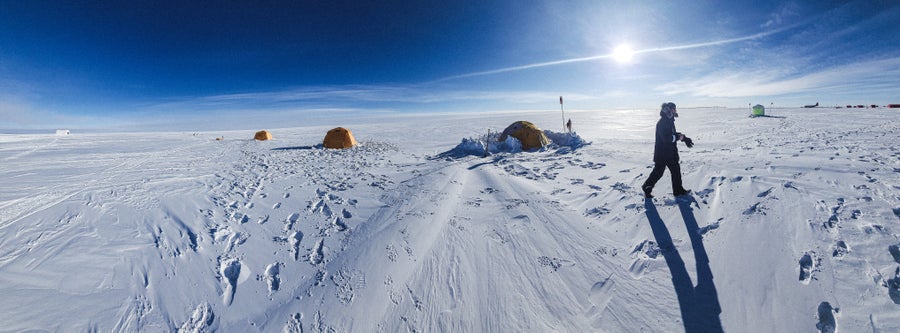 A panoramic view of an ice sheet camp showing several tents and a man walking across the frame with the sun behind him