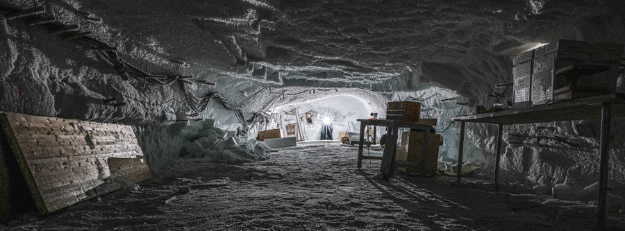 A single light illuminates a low ice cavern featuring several wooden tables topped with tools for measuring and logging ice core samples