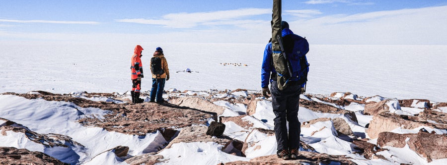 Three people stand looking over the vast expanse of ice on the Greenland ice sheet. Tents can be seen in the distance.
