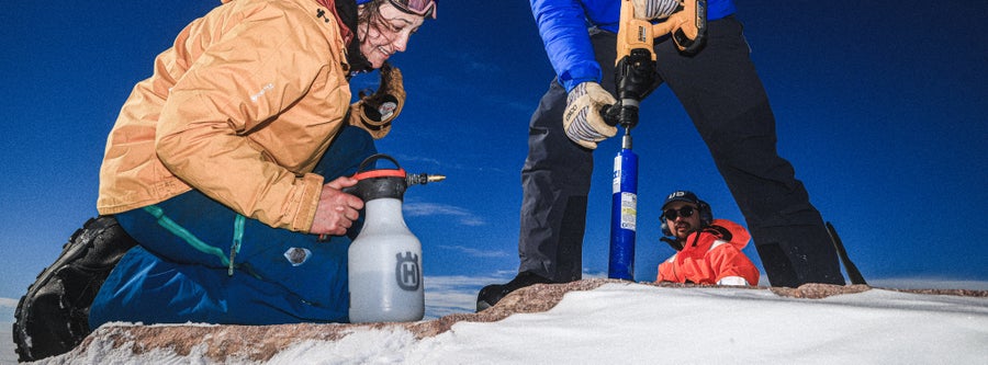 Three people work on a rock against a blue sky. The woman kneels with a spray bottle; one man stands pushing a drill bit into the rock such that we only see his legs and arms; another man peers toward them, seen through the clearing of the other man's spread feet and legs
