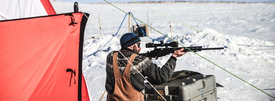 A man in overalls and a thick sweater and cap peers through the scope of a rifle