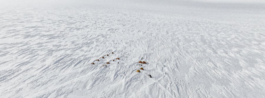 An aerial view of the vast Greenland ice sheet surface with several small, yellow tents dotting its surface in the foreground