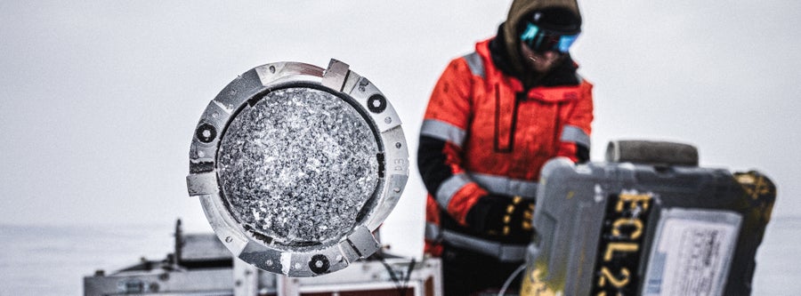 The circular end of an ice drill points toward the camera showing a core barrel full of ice sample and a man standing next to the apparatus in an orange jacket and goggles