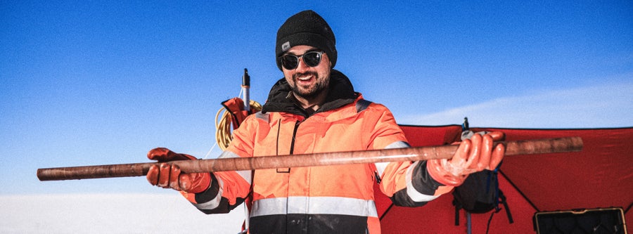 A man in an orange and black jacket with sunglasses and a black snow hat on stands holding a long rock core in his gloved hands
