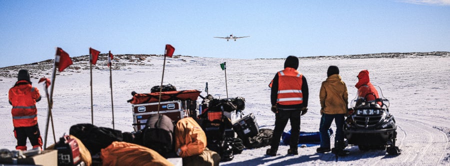 A group of people with their backs to the camera stand in cold weather gear on an ice sheet. They look at a plane with skis that is about to land on an improvised landing strip to their left