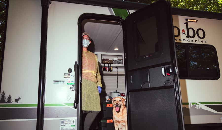 A respiratory masked woman in a laboratory dressing stands at the door or a camper van with a dog