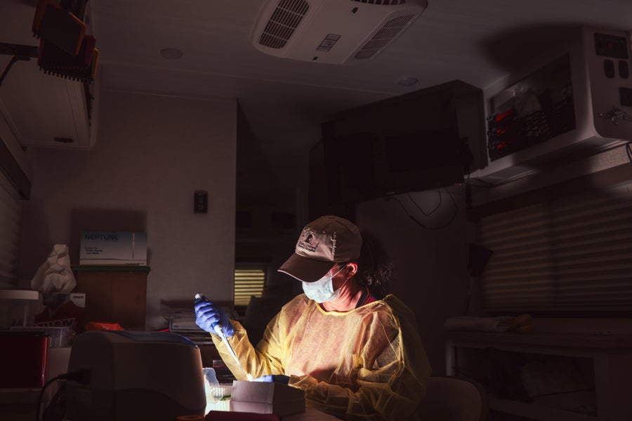 A woman in laboratory scrubs sits in a darkened camper van pipetting.
