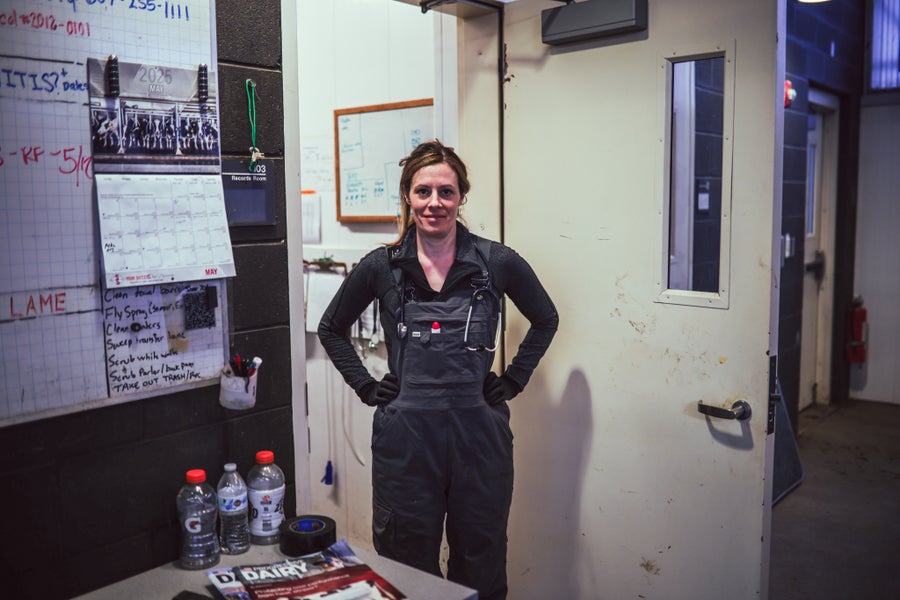 A woman in coveralls stands with hands on hips outside an office door inside a barn