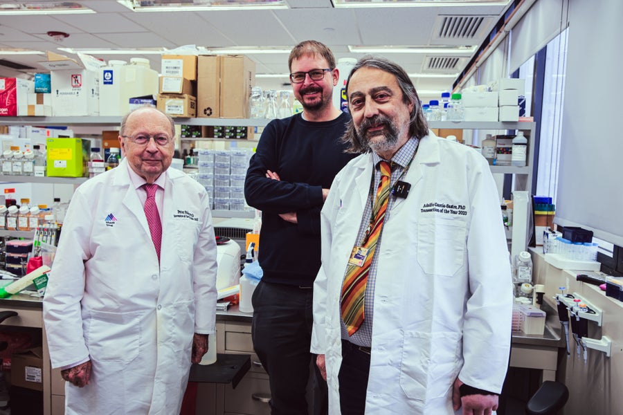 Three men, two in lab coats and one in normal clothing, stand in a research lab facing the camera