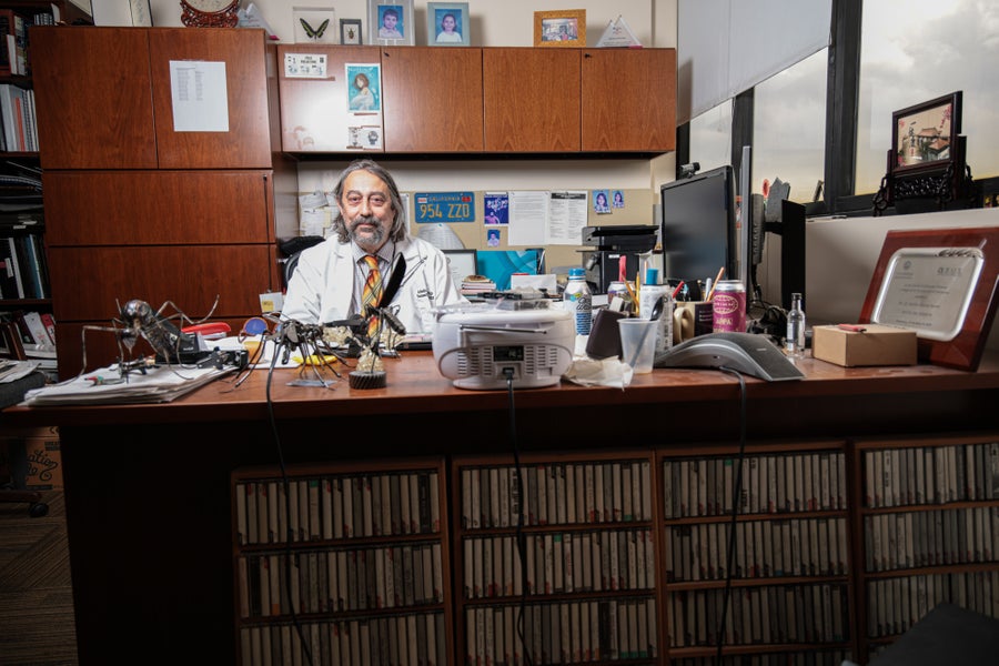 A long-haired older man in a lab coat sits behind a messy desk covered with papers and mosquito statues