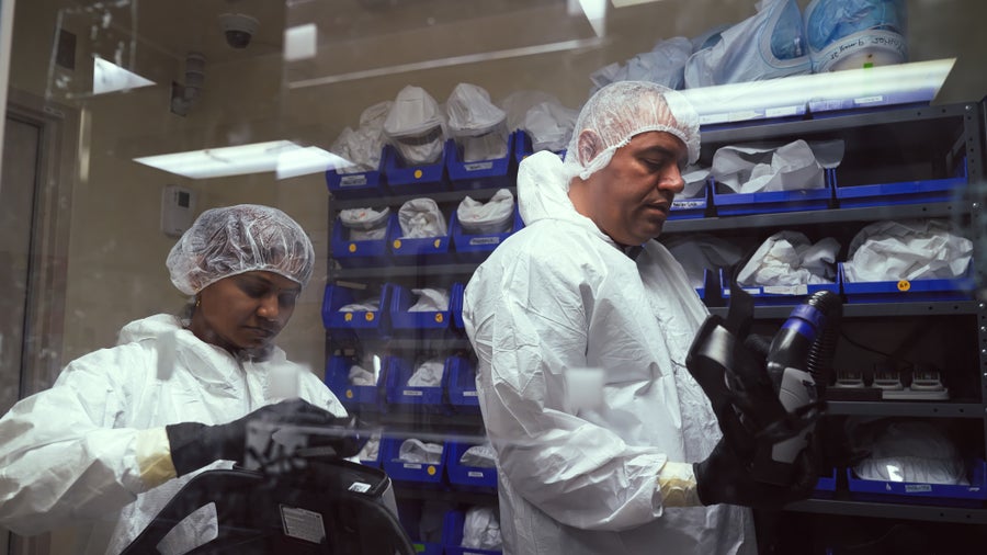 A man and women can be seen dressing up in full body personal protective gear inside a laboratory