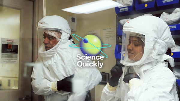 A man and women in full body personal protective gear complete with head shroud and helmet bubble visors are seen inside a laboratory