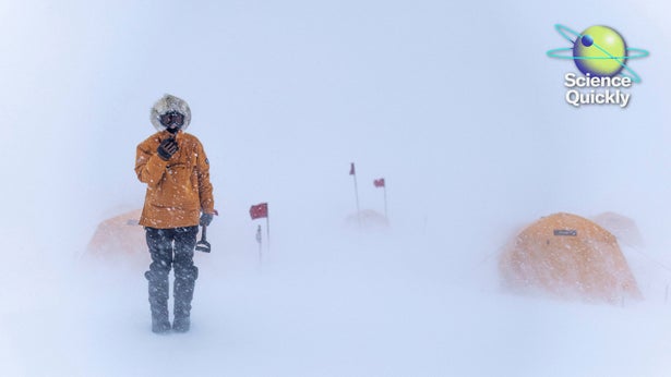 A person in an orange parka stands in a blizzard with red flags and tents barely visible in the near whiteout behind them