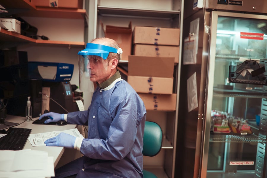 A man in bioharzard personal protective equipment and mask sits at a desk surrounded by sample boxes and lab equipment
