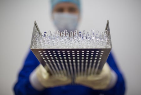 A worker wearing personal protective equipment (PPE) holds a tray containing unlabeled vaccine vials.