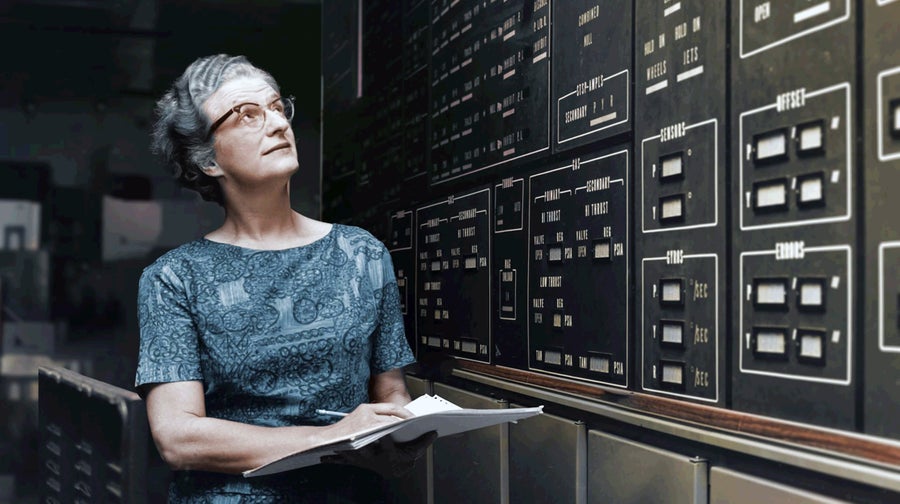 A woman in glasses holds a notebook open as. she leans up and looks at a wall of switches.