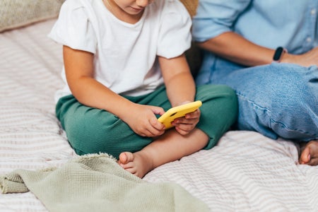 An anonymous child is watching entertainment content on a smartphone with mother sitting next to her on bed