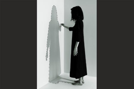 A photo illustration showing a woman facing a wall drawing a dotted line around her shadow.