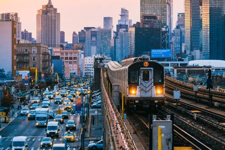 7 Line subway train on an elevated line in Queens with street traffic below to the left and the Manhattan skyline in the background at sunset