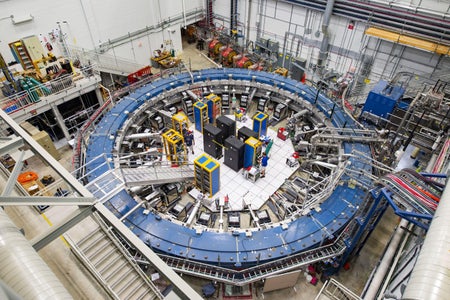 The Muon g−2 ring sits in its detector hall amidst electronics racks, the muon beamline, and other equipment at Fermi National Accelerator Laboratory