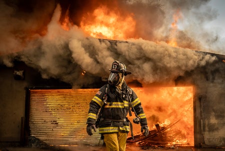 A firefighter in full protective gear walks in front of a building engulfed in flames during the Eaton Fire in Altadena, CA on January 09, 2025