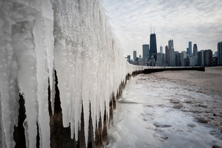 Ice collected along the shore of Lake Michigan with large icicles hanging on the side of a wall and the Chicago skyline in the distance