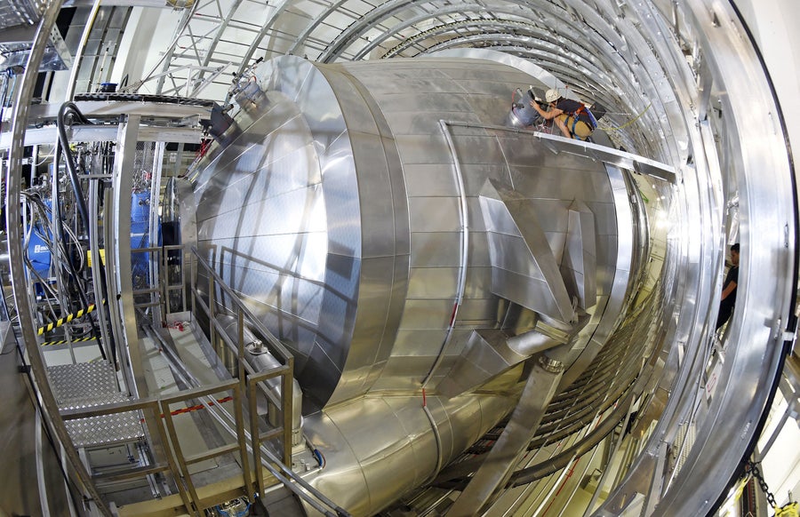 Photo of the main spectrometer of the Karlsruhe Tritium Neutrino Experiment (KATRIN) at the Karlsruhe Institute of Technology (KIT) with a worker, appearing comparatively tiny, kneeling on a platform on the side. The cylindrical giant tank weighs 200 tons, with a length of 24 meters, a diameter of 10 meters and an inner surface of 800 square meters.
