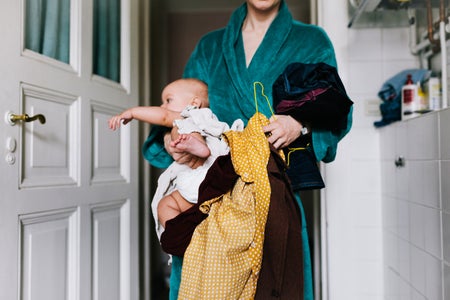 Mother in bathroom wearing a green robe, struggling to carry a baby, clothing on a hanger and in crux of arm simultaneously