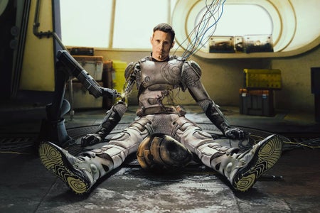 A blend of a robot and man sitting on the floor with legs outstretched and looking at the camera with a plain look.