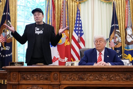 Elon Musk (left) stands and shrugs while speaking to reporters alongside U.S. President Donald Trump (right), sitting at the Resolute Desk, in the Oval Office of the White House on May 30, 2025