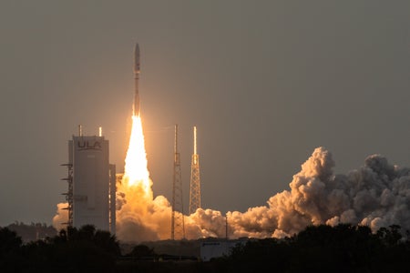 A rocket launches at night in front of a structure labeled ULA