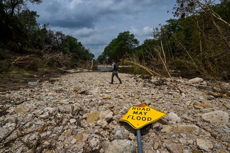 A member of search and rescue personell walks along Guadalupe River in Texas after a devastating flash flood. In the foreground, a toppled sign reads, "ROAD MAY FLOOD."