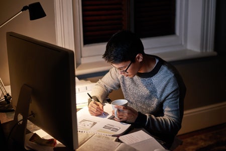 Young college student pulling an all nighter, studying papers on the desk in front of a computer screen, holding coffee and a pen, illuminated by a single desk lamp