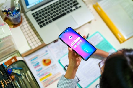 Student interacts with an AI chatbot on a smartphone while studying at a desk with a laptop, notes and stationery.