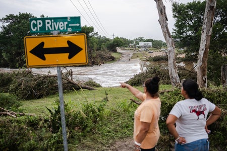 Two women in the foreground standing on a roadside survey the damage caused by flash flooding on the bank Guadalupe River on July 5, 2025 in Center Point, Texas