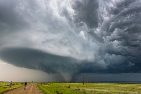 Storm chasers document a tornado beneath a dramatic supercell storm along a rural dirt road in scenic Montana