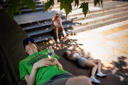 In the foreground, a woman in a green shirt lies on wall in the shade with a bottle of water in her hands. In the background, a teen girl is lying on her side in the shade and another woman sits on the stairs behind her