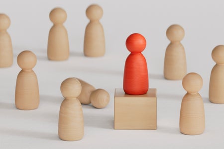 Photo illustration, wooden pawn figures gathered around a red pawn figure standing on a pedestal. One wooden pawn figure is overturned laying on the floor.