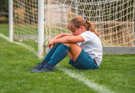 Side view of tired 9 year old female soccer player sitting on field next to goal thinking about how she played in practice.