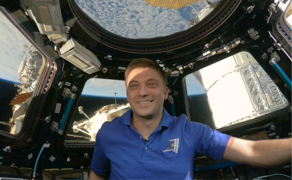 Matthew Dominick (a white man with short dirty blonde hair) smiles in front of the windows of the cupola, through which you can see Earth. He is wearing a bright blue t-shirt that matches the blue of the Earth behind him.