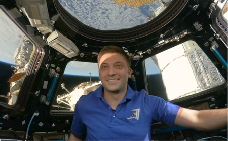 Matthew Dominick (a white man with short dirty blonde hair) smiles in front of the windows of the cupola, through which you can see Earth. He is wearing a bright blue t-shirt that matches the blue of the Earth behind him.