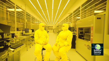 Two people in full clean room bunny suits sit on stools facing each other in a large yellow-colored room